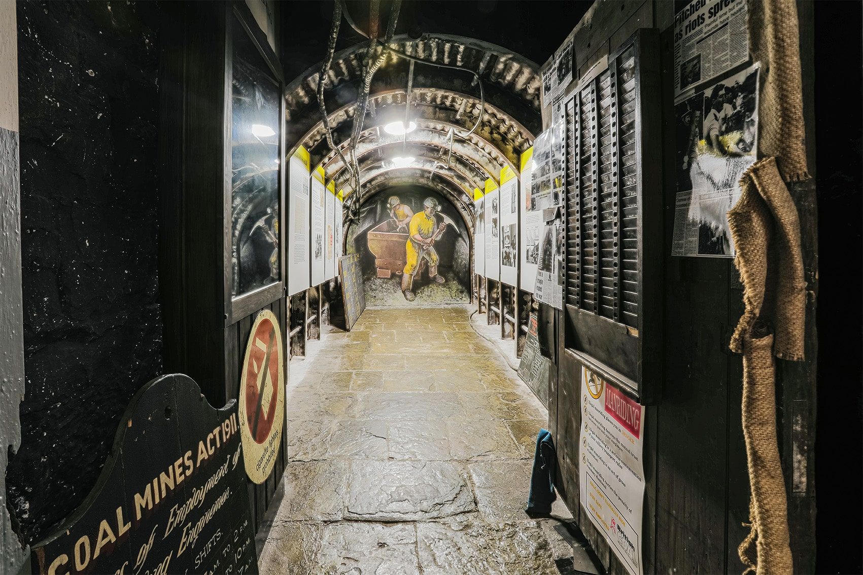 A narrow dark passageway with a paved floor, information panels down either side and an illustration of a miner at the end.jpg