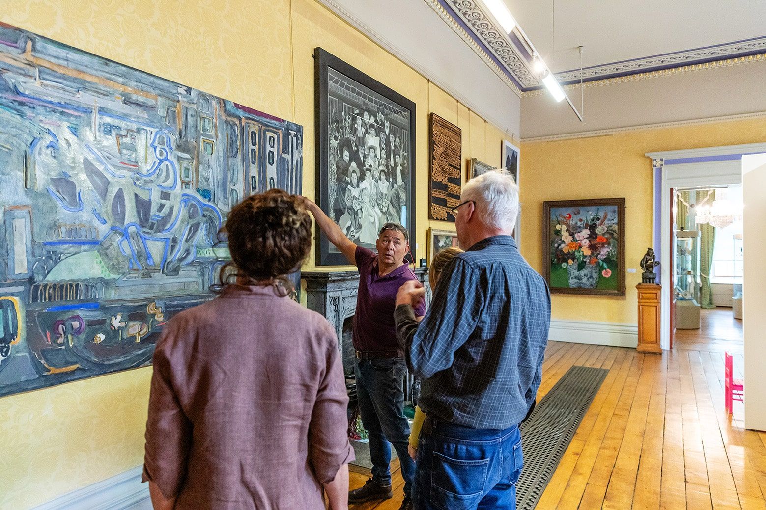 A man pointing to a modern painting hanging on a yellow wall with people stood around watching him.jpg