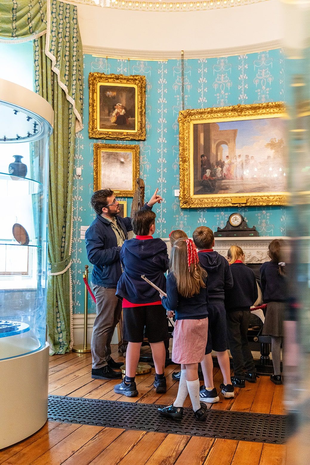 Children in navy and red uniform looking at paintings in gold frames hung on a wall with turquoise wallpaper.jpg